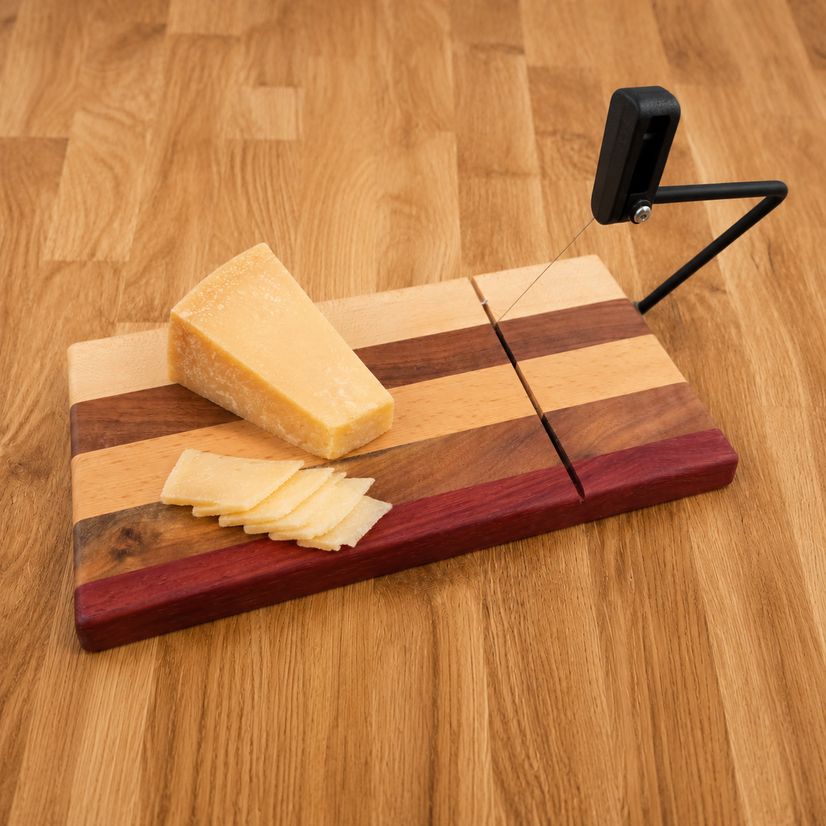 A striped wooden cheese board with a black crank-style cheese slicer on top, shown with a wedge of cheese and several slices on a wood-grain tabletop.