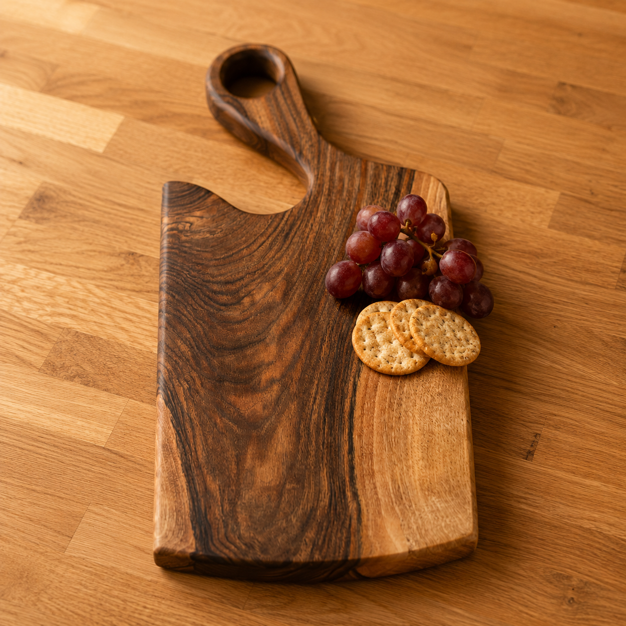 A dark wood charcuterie board with a handle, styled on a light wood table with red grapes and two crackers.