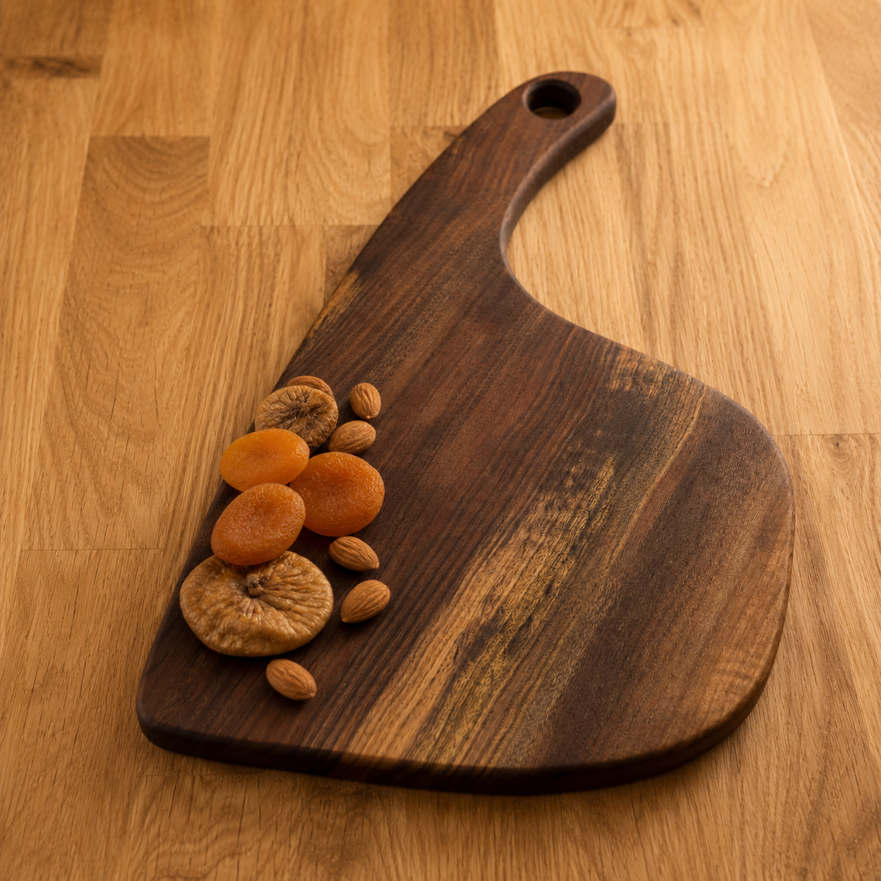 A dark brown walnut serving board with a handle, styled with dried apricots and almonds on a light wood tabletop.