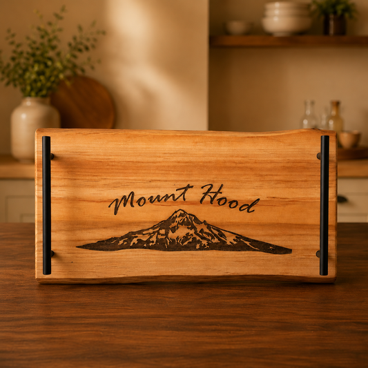 A rectangular wooden serving tray with live-edge natural wood sides and black metal handles, featuring a dark engraved mountain graphic and the words 'Mount Hood' on the front, set on a wooden table in a warm, softly blurred kitchen interior.