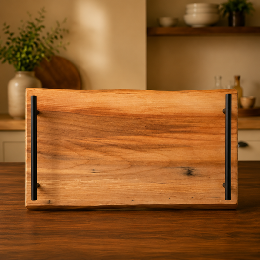 A rectangular wooden serving tray with a live edge and two black metal handles, shown on a wooden table in a warm, softly blurred kitchen setting.
