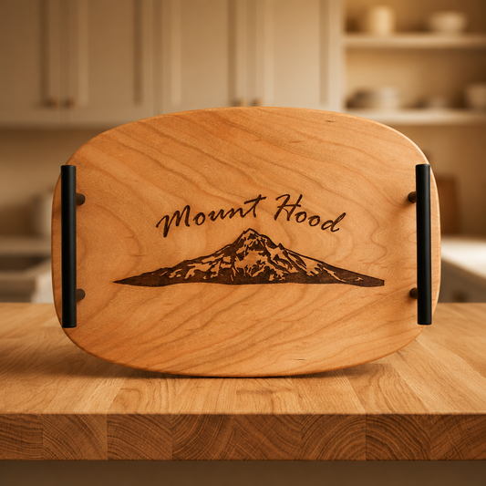 A round wooden serving board with black handles and an engraved 'Mount Hood' mountain design, displayed on a wooden countertop with a blurred kitchen background.