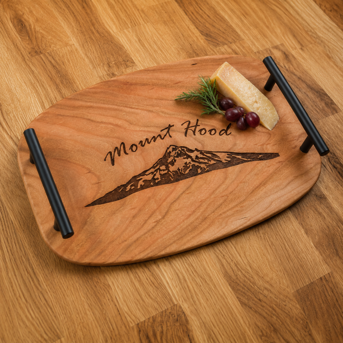 A wooden serving tray with black handles, engraved with 'Mount Hood' and a mountain graphic, styled with cheese, grapes, and rosemary on a wood floor.