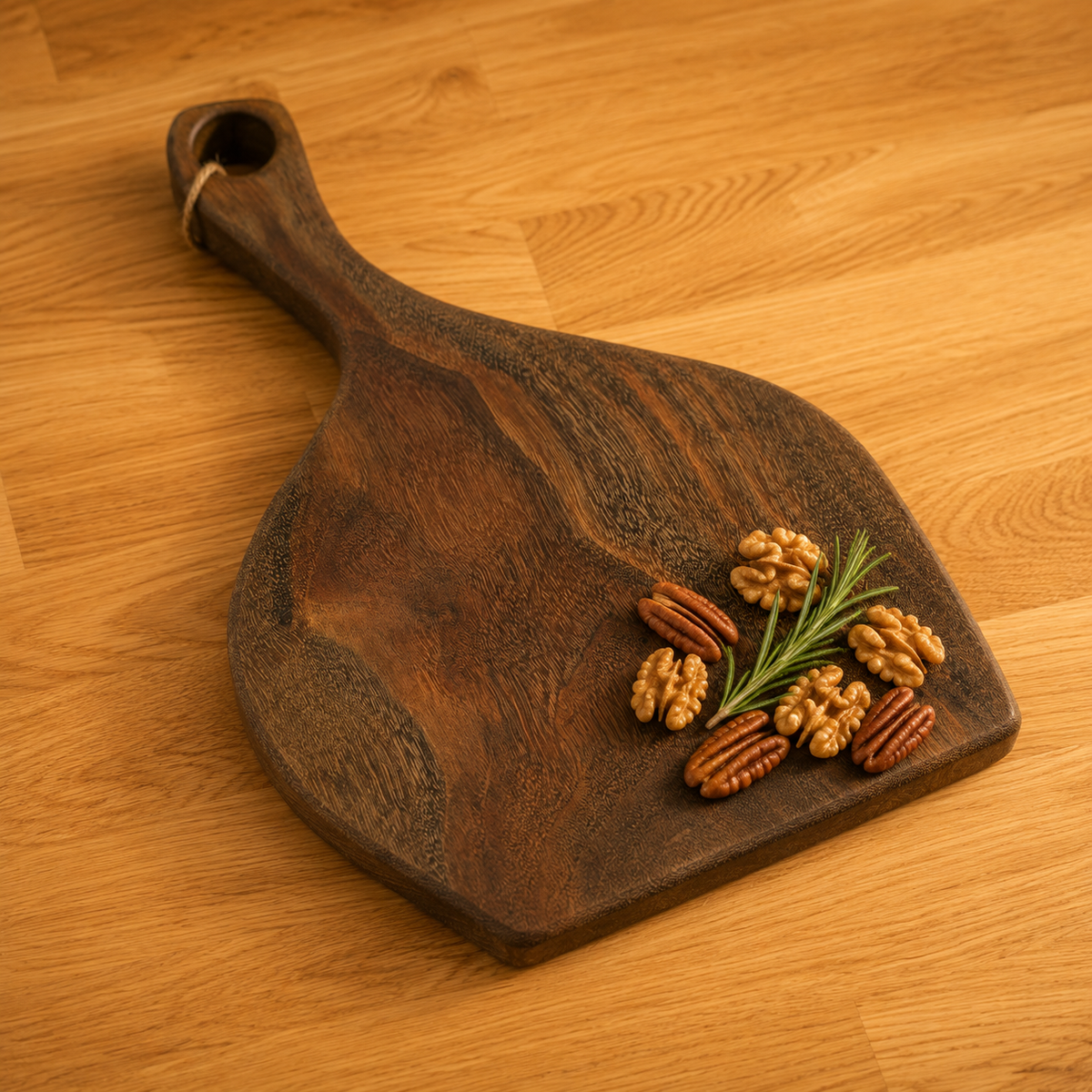 A dark brown walnut wooden serving board with a handle, styled with walnuts and a sprig of rosemary on a light wood tabletop.
