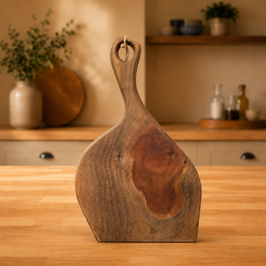 A rustic walnut charcuterie board with a handle and a natural wood grain pattern, standing on a wooden countertop in a warm, softly lit kitchen setting.