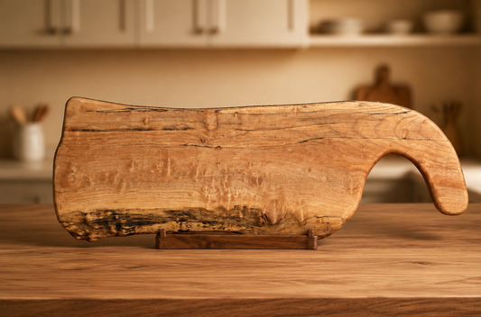 A rustic, maple-handcrafted wooden charcuterie board with a natural live edge and a curved handle, displayed on a wooden table in a warm, softly blurred kitchen setting.
