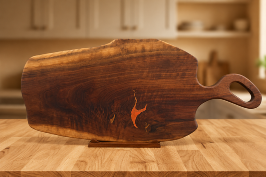A rustic walnut charcuterie board with a live edge and a loop handle, displayed on a wooden countertop with a blurred kitchen background.