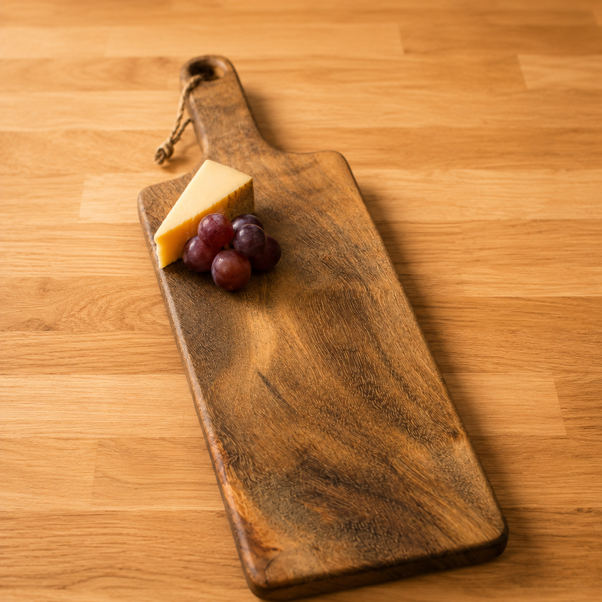 A rustic dark wood cheese board with a handle and hanging loop, styled on a light wood tabletop with a wedge of cheese and red grapes on top.