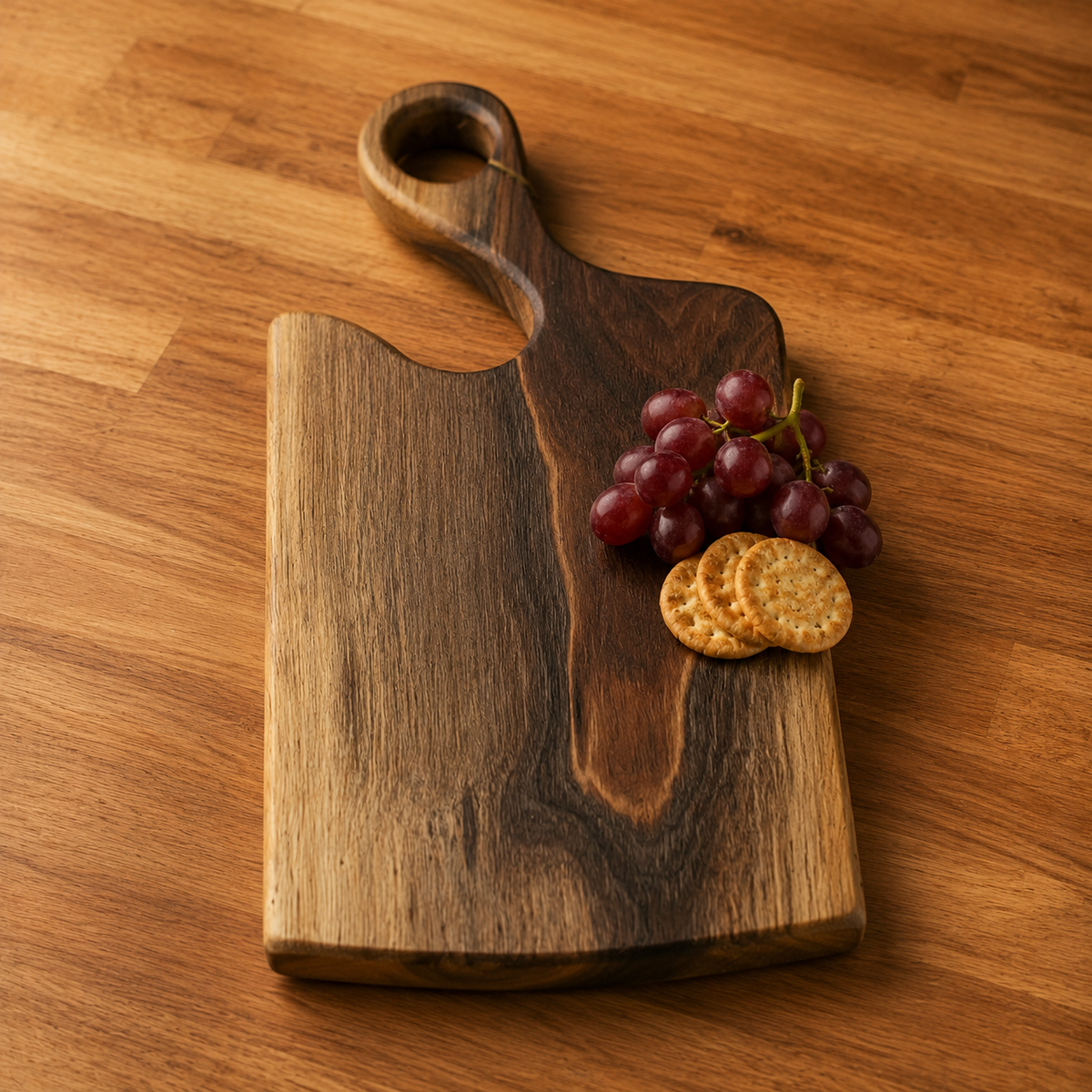 A rectangular wooden charcuterie board with a rounded handle and a natural wood grain pattern, styled on a wooden table with red grapes and round crackers.