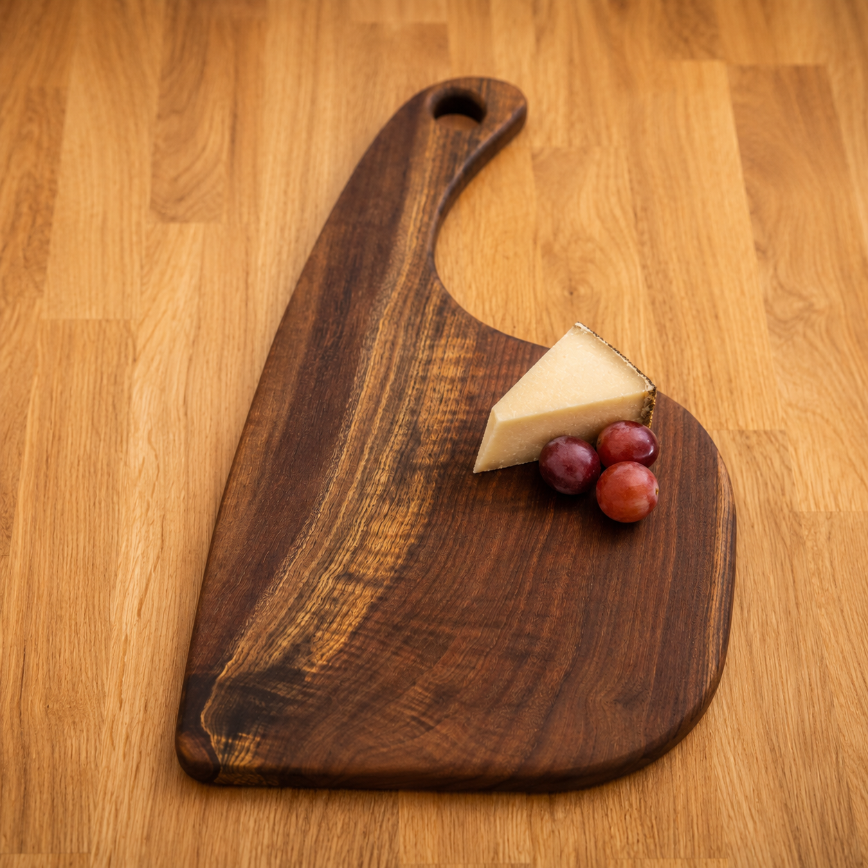 A dark brown walnut serving board with a handle, styled with a wedge of cheese and three red grapes on a light wood tabletop.