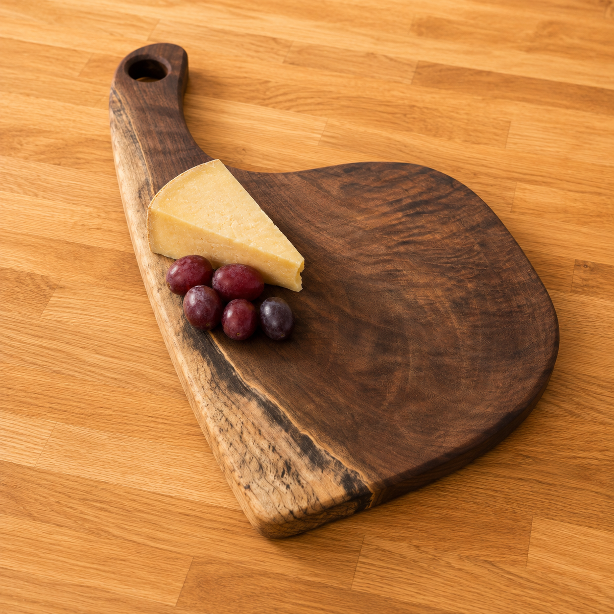A rustic walnut serving board with a handle, styled on a light wood table with a wedge of cheese and three purple grapes.