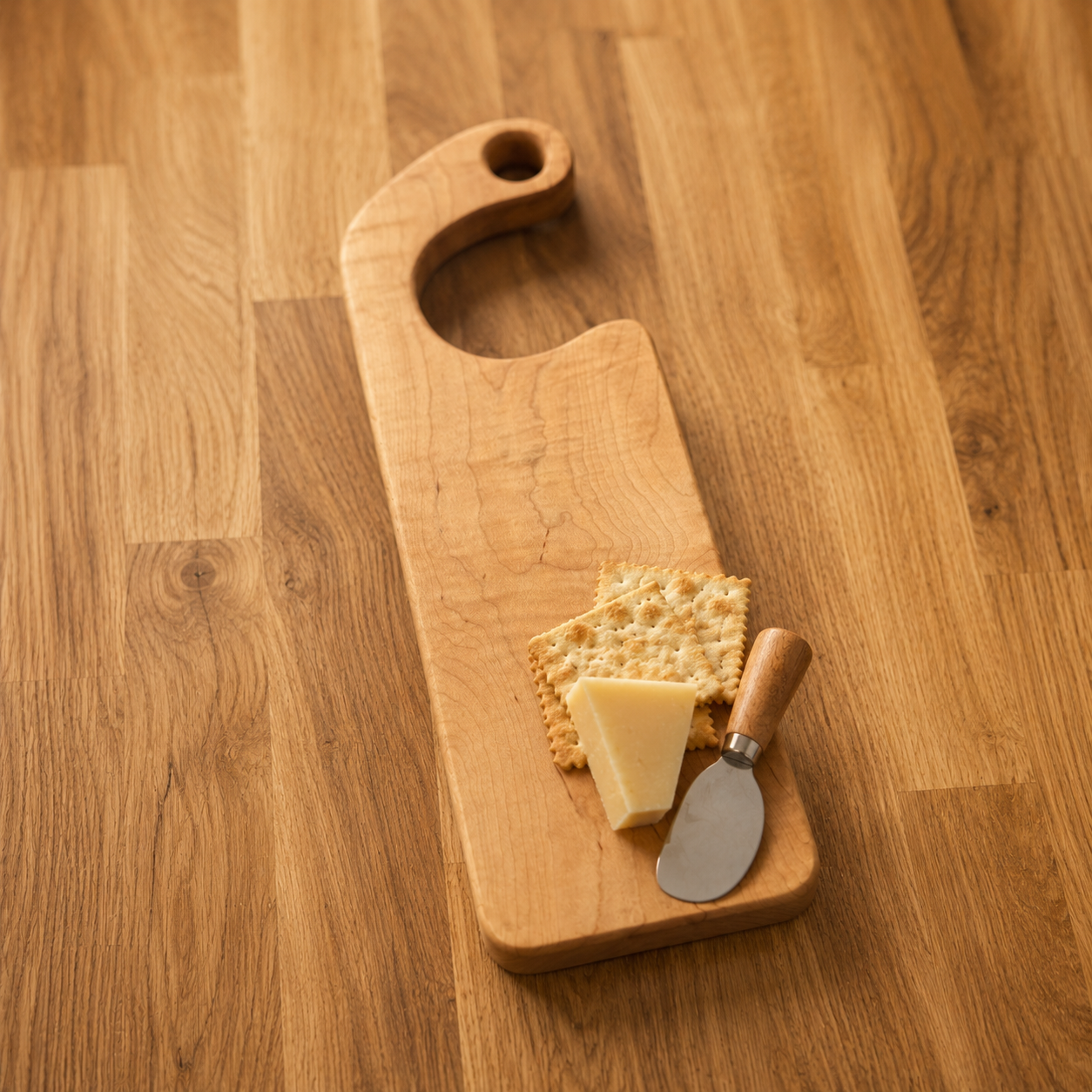 A rectangular wooden serving board with a curved handle and a round cutout, shown on a wood countertop with a cracker, cheese wedge, and small knife placed on top.
